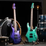 Two electric guitars stand upright on a stage beside a blue sparkle drum set in the background, one purple-blue and one teal-green. ขายราคาพิเศษ