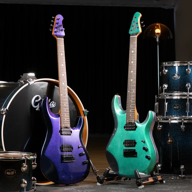 Two electric guitars stand upright on a stage beside a blue sparkle drum set in the background, one purple-blue and one teal-green. ขายราคาพิเศษ