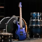 Blue electric guitar with a shimmering finish rests against a large black bass drum in a drum set arrangement on a dark stage backdrop. ขายราคาพิเศษ