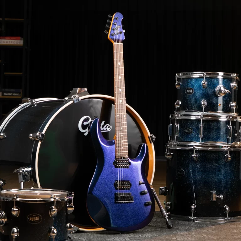 Blue electric guitar with a shimmering finish rests against a large black bass drum in a drum set arrangement on a dark stage backdrop. ขายราคาพิเศษ
