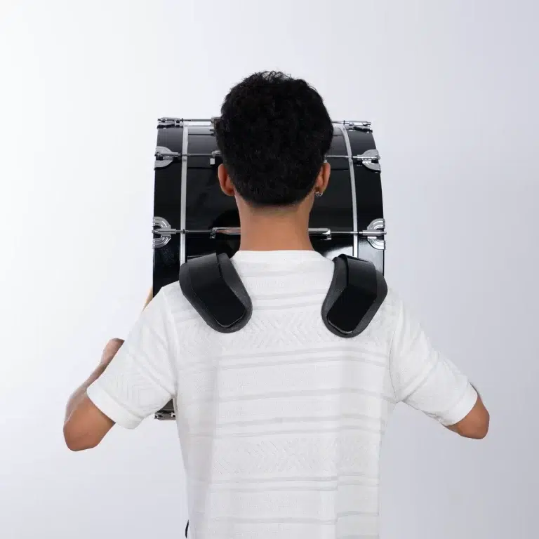Child seen from behind carrying a large black bass drum with metal lugs, strapped to a harness ขายราคาพิเศษ