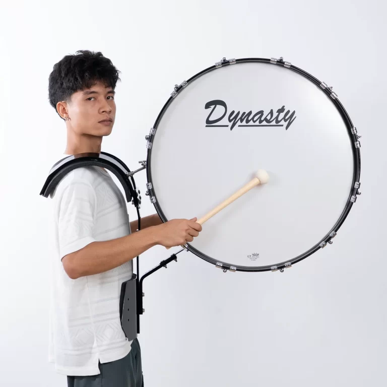 Young man wearing a white shirt plays a large bass drum on a harness, striking it with a wooden mallet; drumhead reads 'Dynasty'. ขายราคาพิเศษ
