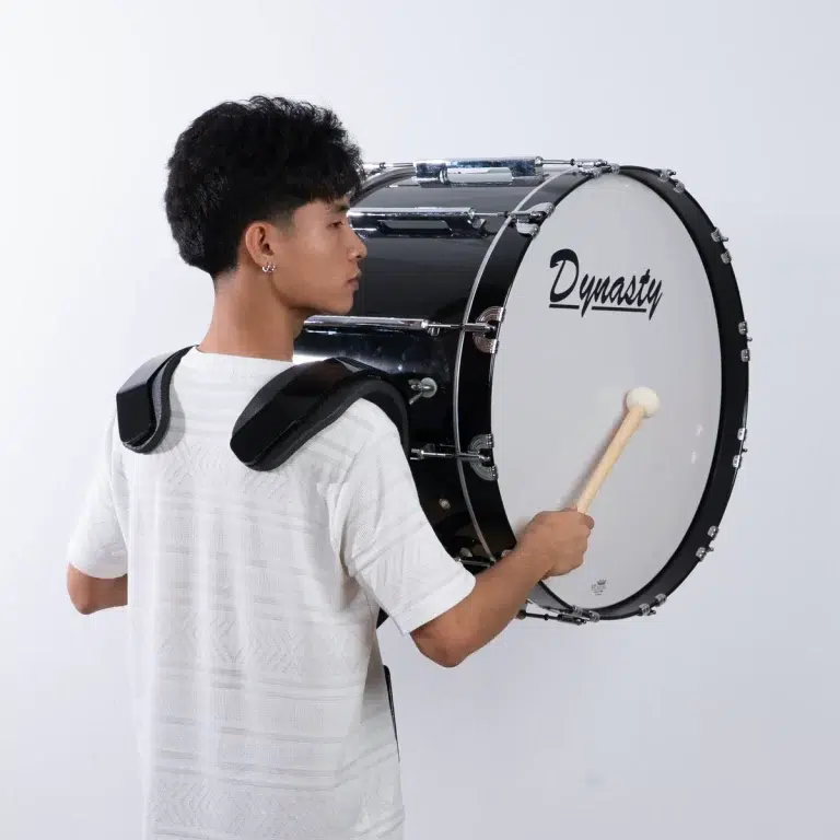Drummer in a white shirt wearing a shoulder harness carries a large Dynasty bass drum with a mallet resting on the drumhead. ขายราคาพิเศษ
