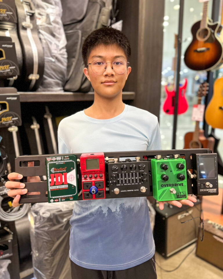Young person with glasses in a music shop holding a multi-pedal guitar effects pedalboard
