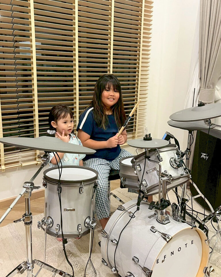 Two young girls sit behind a white drum kit indoors, holding drumsticks and posing by the blinds.