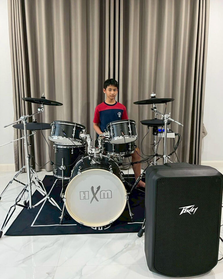 Young boy sits behind a black drum kit in a room with gray curtains, practicing.