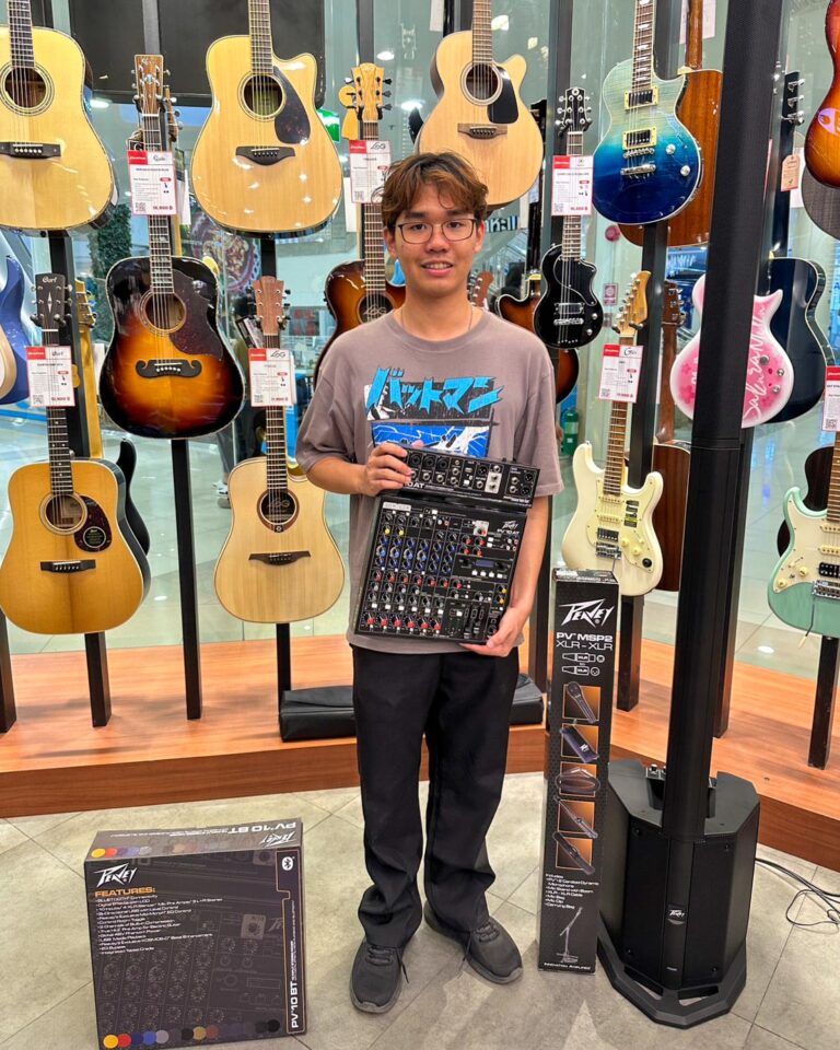 Young man in a guitar shop holds a compact mixer and stands among guitar displays with price tags visible.