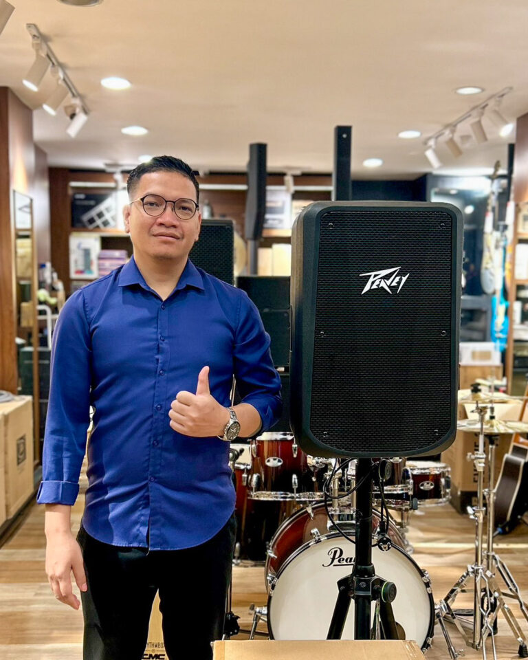 Man in a blue shirt and glasses gives a thumbs up beside a large black Peavey speaker in a music store, with drums visible in the background.