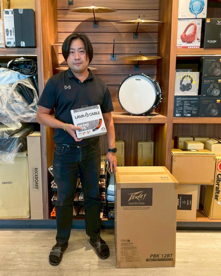 Man standing in a music store holding a Lava Cable box, with drum gear and cymbals on wooden shelves behind him.