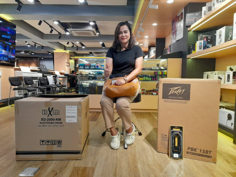 Woman seated on a stool in an electronics store, giving a thumbs up, with large boxes for a drum set and accessories nearby.