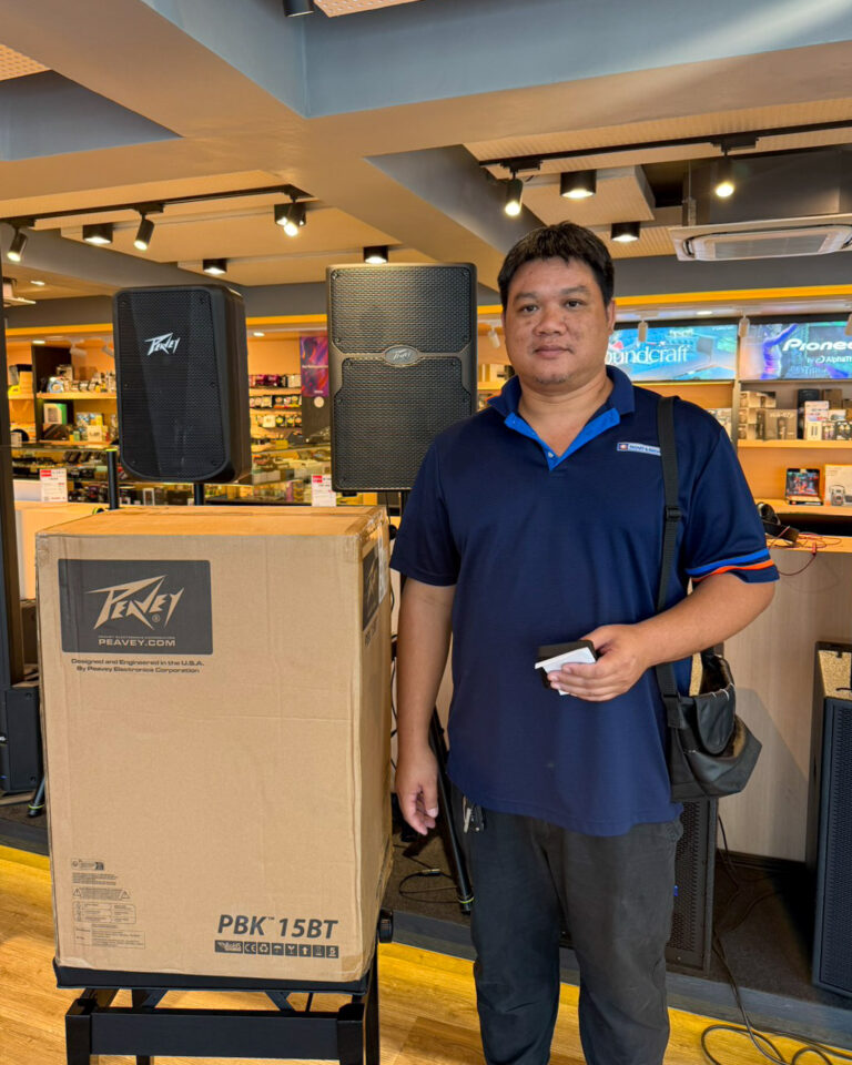 Man in a blue polo stands next to a large speaker box on a display in an electronics store, with two tall PA speakers visible behind him.