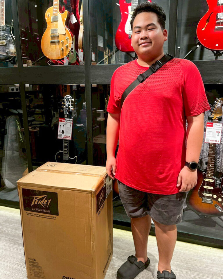 Person in a red shirt standing beside a large Peavey box in a guitar shop with guitars on display behind him.