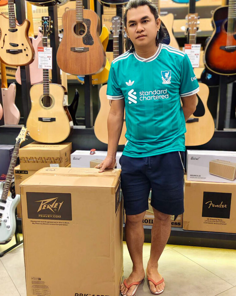 Young man in a turquoise jersey stands in a guitar shop, resting his hand on a large Peavey box among hanging guitars and other boxed gear.
