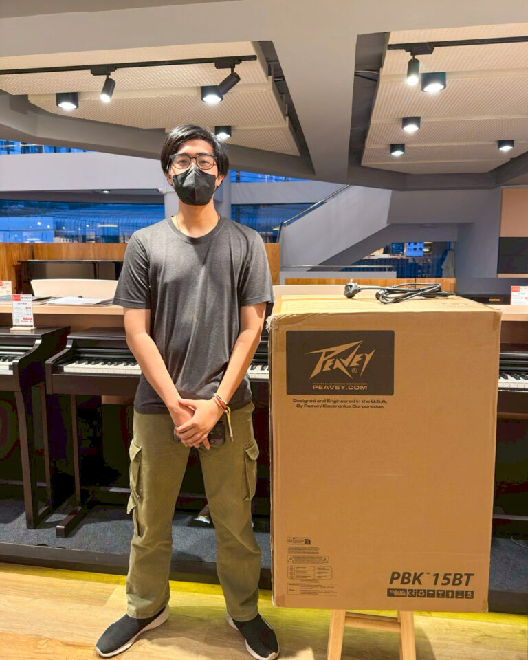 A person wearing a black face mask and glasses stands in a showroom beside a large cardboard box labeled PBK 15BT, with keyboards in the background.