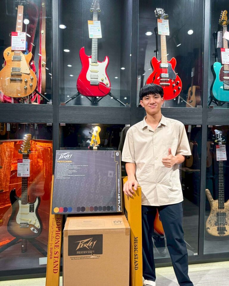 Smiling young man stands beside a glass guitar display in a music shop with colorful electric guitars behind him, giving a thumbs up.