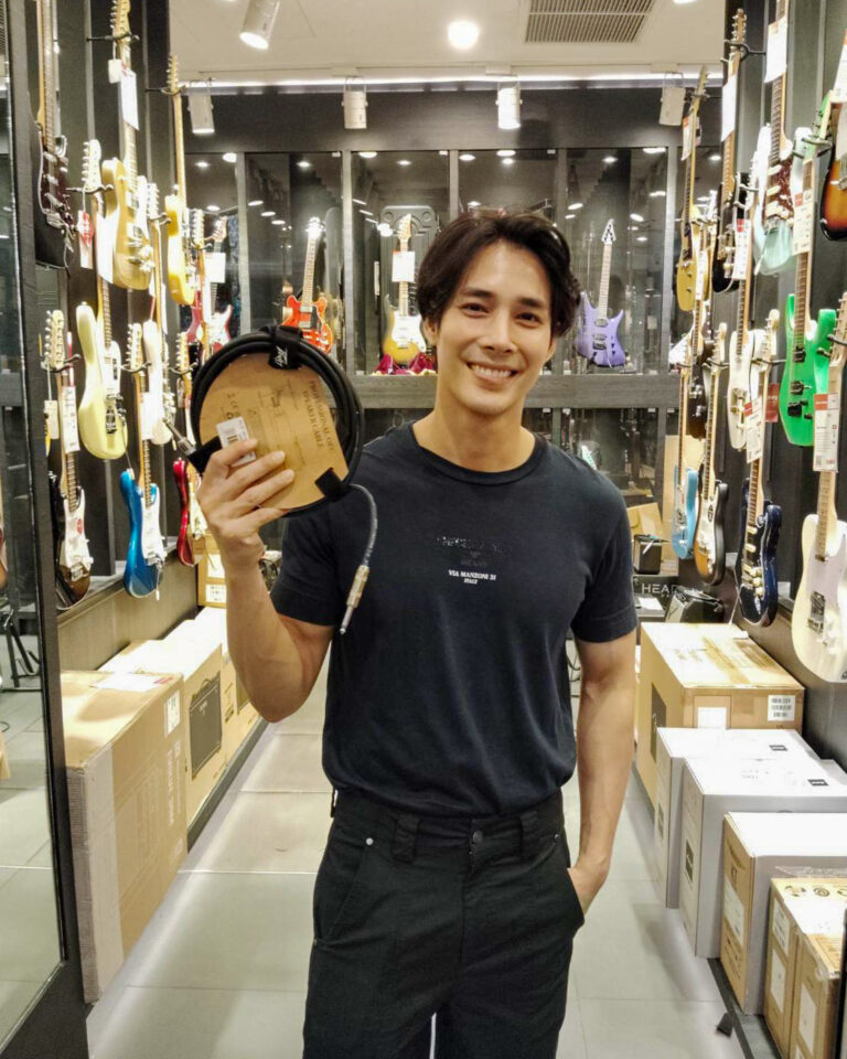 Smiling man in a guitar shop holds a coiled pair of headphones in front of guitar display with various guitars on the walls behind him.