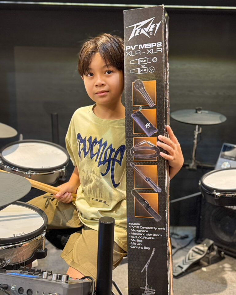 Young boy sits at a drum set in a music room, holding a tall Pearl box of microphone and accessory kit beside him.
