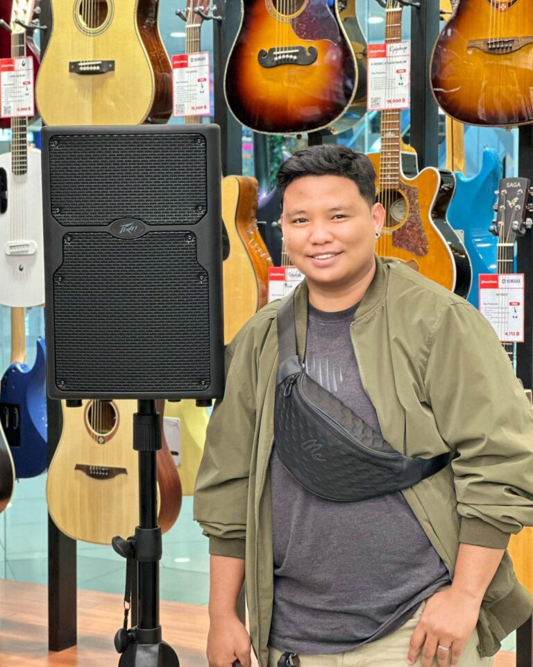 Man posing in a guitar shop with acoustic guitars on display behind him and a large black speaker on a stand in the foreground, smiling at the camera.