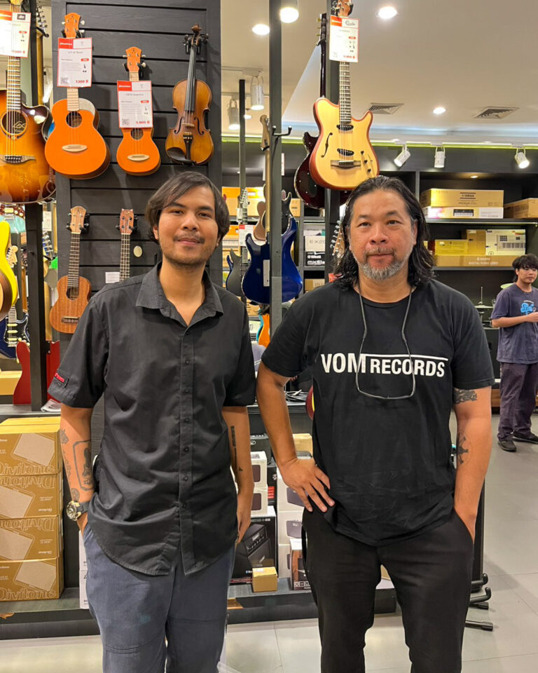 Two men stand in a music store in front of guitar displays, one wearing a black shirt and the other a VOM Records tee.