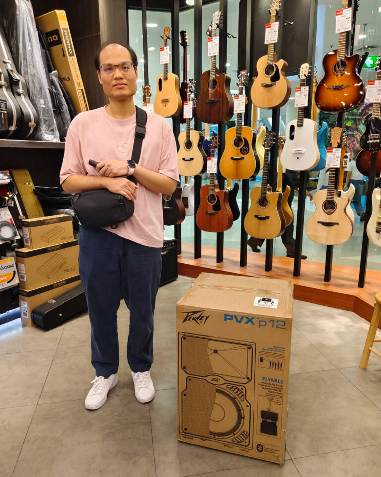Man wearing a pink shirt and glasses posing in a guitar shop, with numerous acoustic guitars hanging behind him and a large PVX p12 amp box on the floor in front.