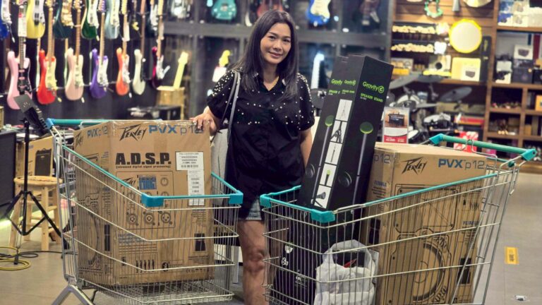 Woman in a music store pushing a shopping cart filled with large boxed gear, guitar displays in the background.