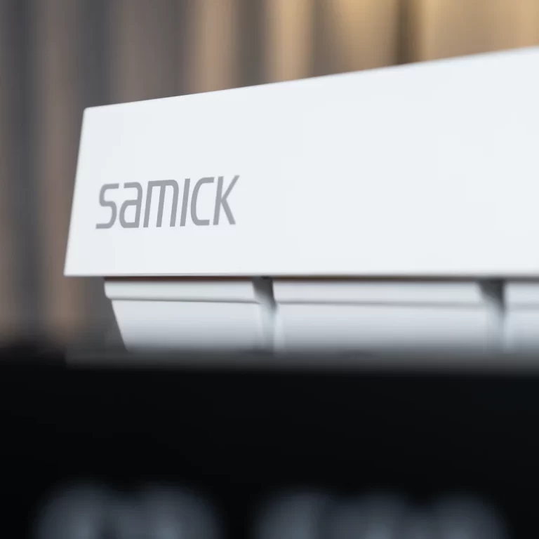 White angled display sign with the word 'samick' in gray lettering against a blurred background. ขายราคาพิเศษ