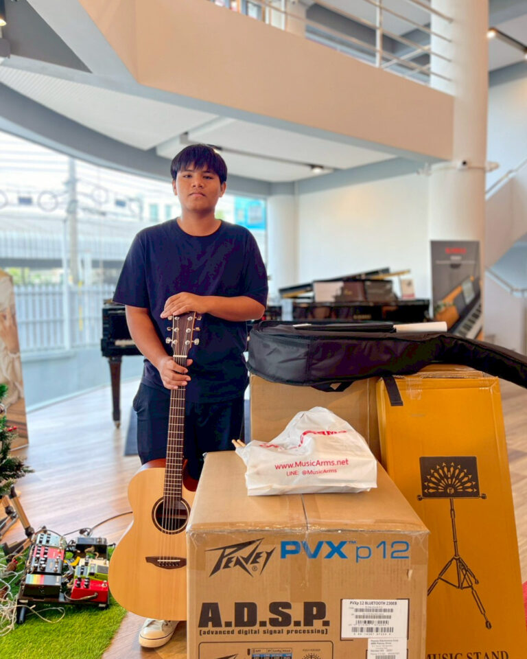 Young man stands in a music store holding an acoustic guitar, surrounded by boxes and a grand piano in the background.