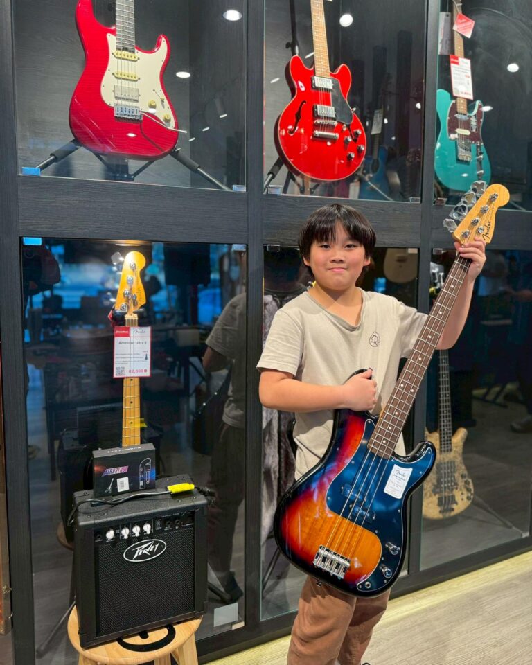 Young boy posing with a sunburst Fender bass guitar in a guitar shop window display with red and teal guitars in the background.