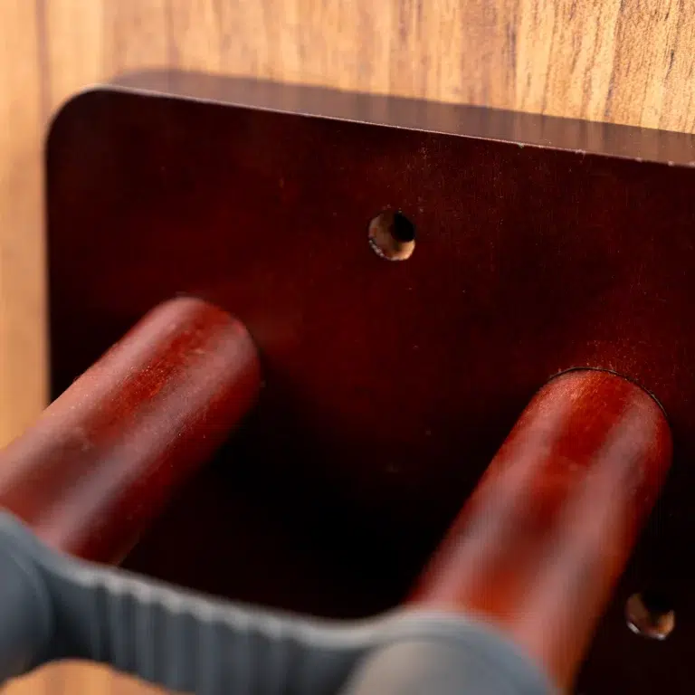 Close-up of a dark brown wooden panel with a drilled hole and two wooden dowel-like rods nearby, held by a gloved hand. ขายราคาพิเศษ