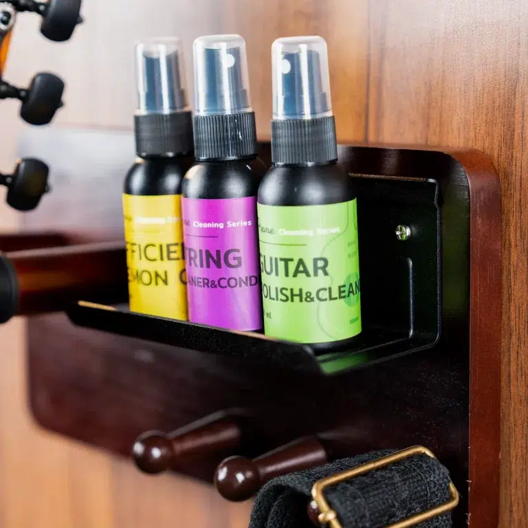 Three spray bottles with yellow, purple, and green labels reading 'GUITAR POLISH & CLEAN' sit on a dark wooden shelf; a strap and buckle are visible below. ขายราคาพิเศษ