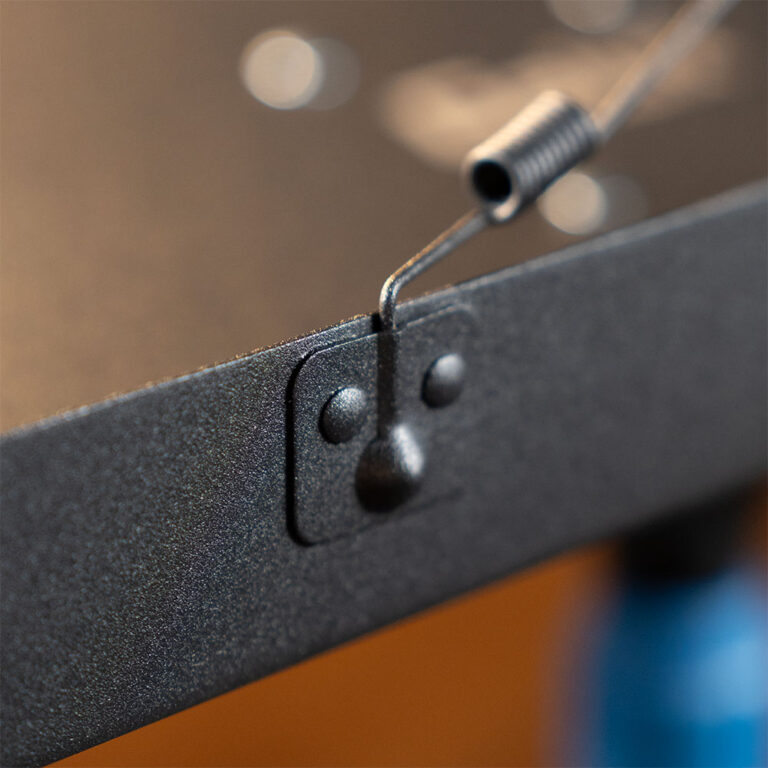 Macro shot of a dark metal frame with a curved wire pin securing a hinge-like latch. The coiled end of the pin is visible against a textured panel. ขายราคาพิเศษ