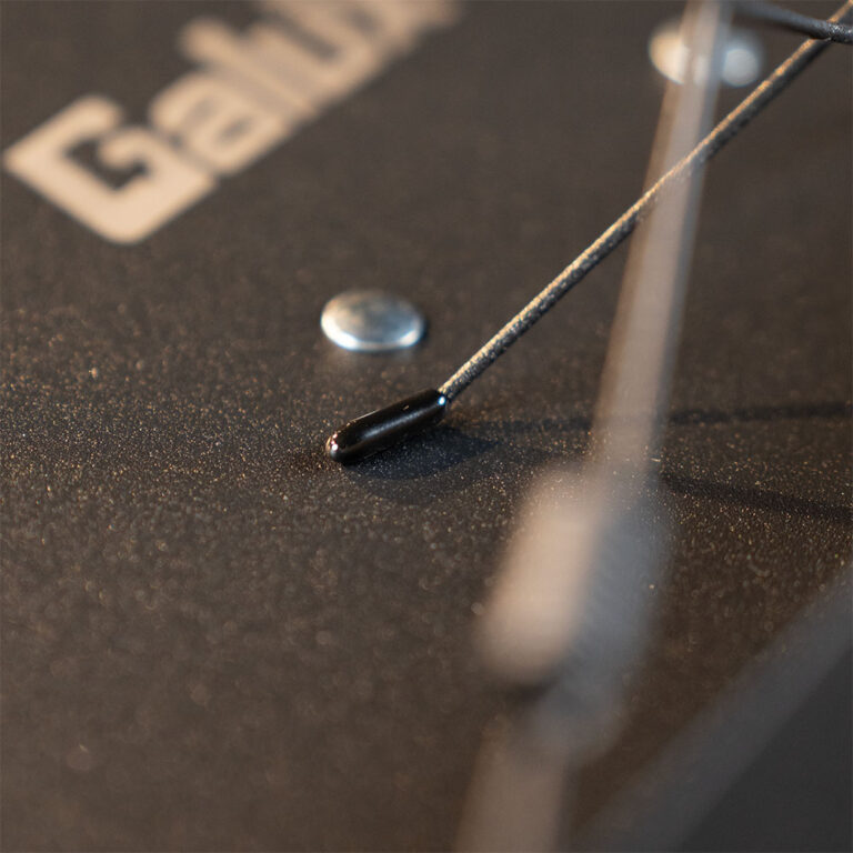 Close-up of a thin wire or probe with a small black tip resting on a dark textured surface, with a silver round object nearby. ขายราคาพิเศษ