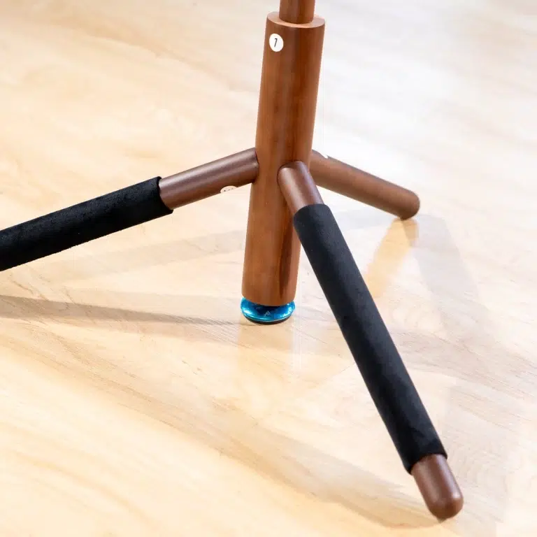 Wooden tripod-like stand with three foam-wrapped legs splayed on a light hardwood floor; a blue circular foot is visible beneath one leg. ขายราคาพิเศษ