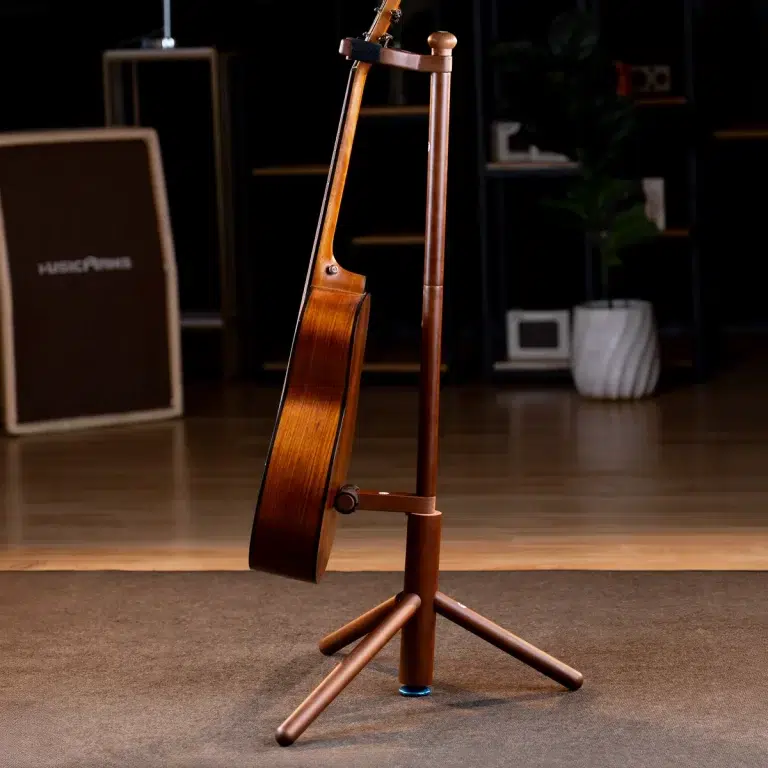 A wooden acoustic guitar resting on a stand in a dimly lit rehearsal space, with an amp and plant in the background. ขายราคาพิเศษ
