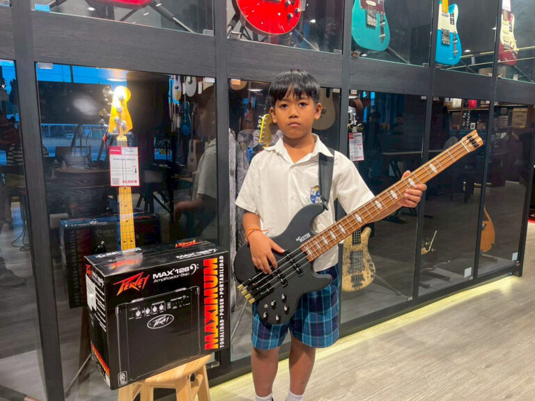 Boy in white shirt and plaid shorts holds a black bass guitar in a music store, with guitars on wall behind him and an amplifier box nearby.