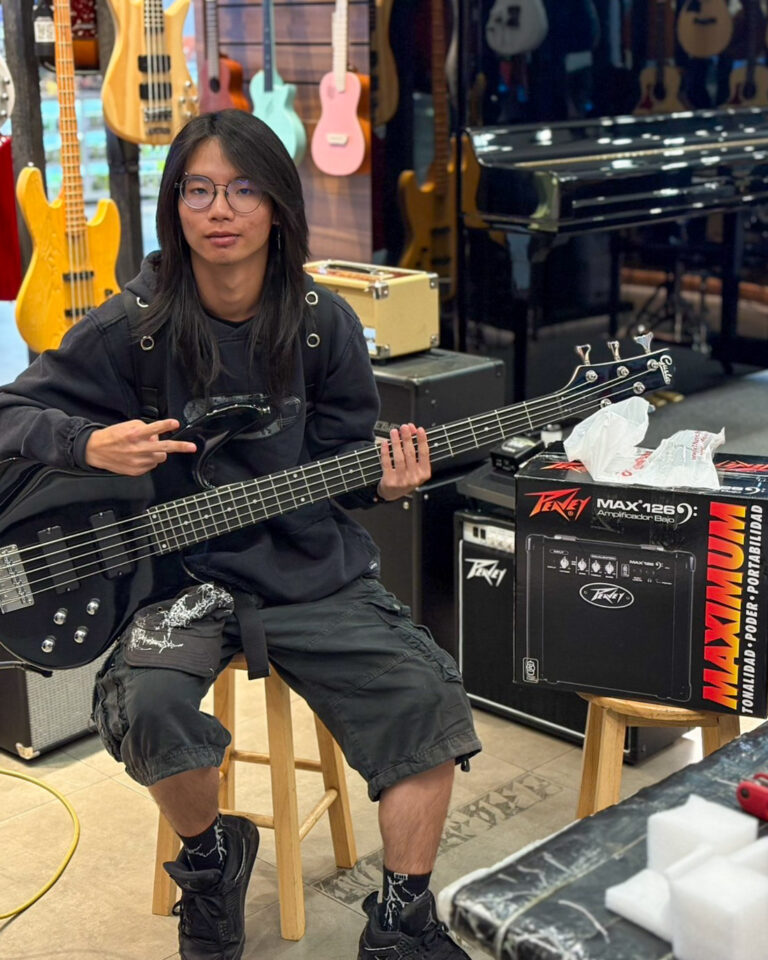 Person sits on a stool in a guitar shop, playing a black five-string bass with colorful guitars hanging in the background.