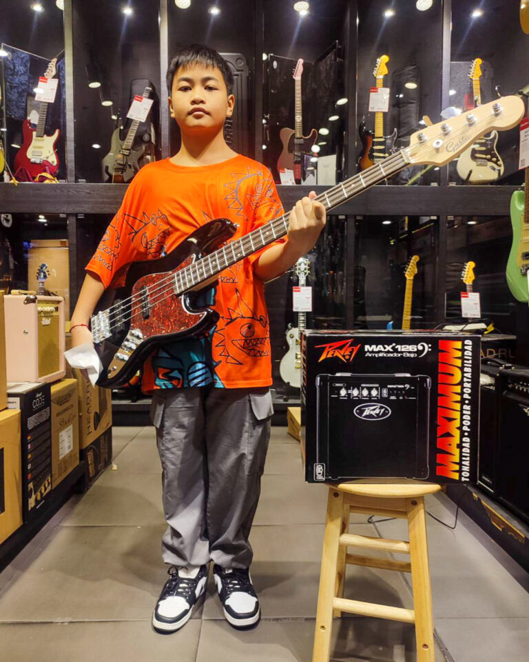 Young boy in an orange shirt stands in a guitar shop, holding a black bass guitar with a red pickguard.