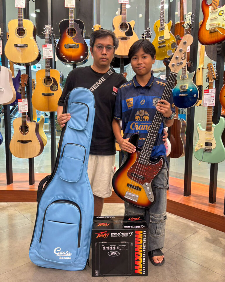 Two people pose in a guitar shop surrounded by hanging guitars; one holds a sunburst bass guitar, the other a blue gig bag, with amplifier boxes on the floor nearby.