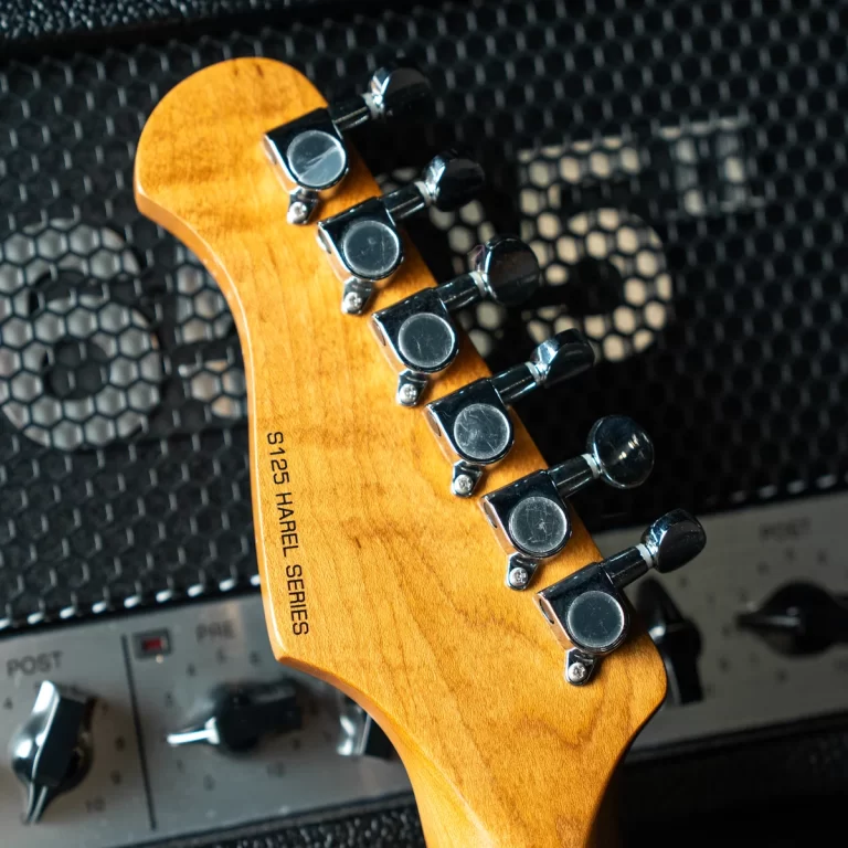 Close-up of a wooden guitar headstock with chrome tuning pegs, against a textured amplifier background. ขายราคาพิเศษ