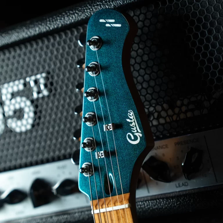 Close-up of a teal electric guitar headstock with six tuning pegs and strings, displaying the Gibson logo, resting against a black amplifier grille behind. ขายราคาพิเศษ