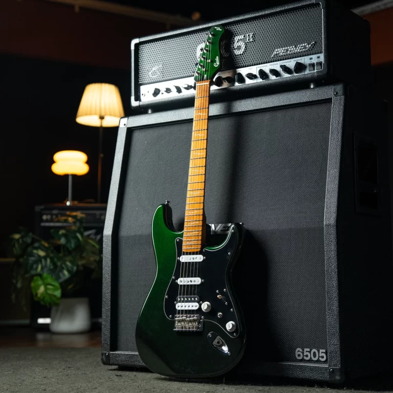 Green electric guitar with a maple neck leaning against a tall black guitar amplifier stack in a dimly lit room, a warm floor lamp glowing in the background. ขายราคาพิเศษ