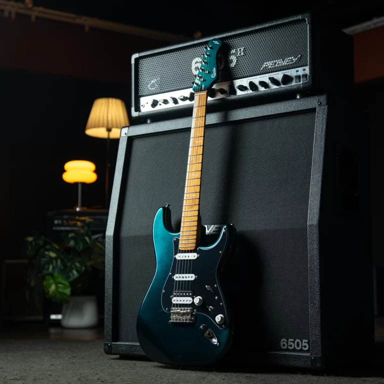 Teal electric guitar leaning against a large black amplifier stack in a dimly lit room, with warm lamps in the background. ขายราคาพิเศษ