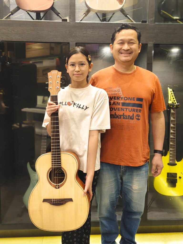 Girl in a white shirt holding an acoustic guitar beside a smiling man in an orange shirt inside a guitar shop.
