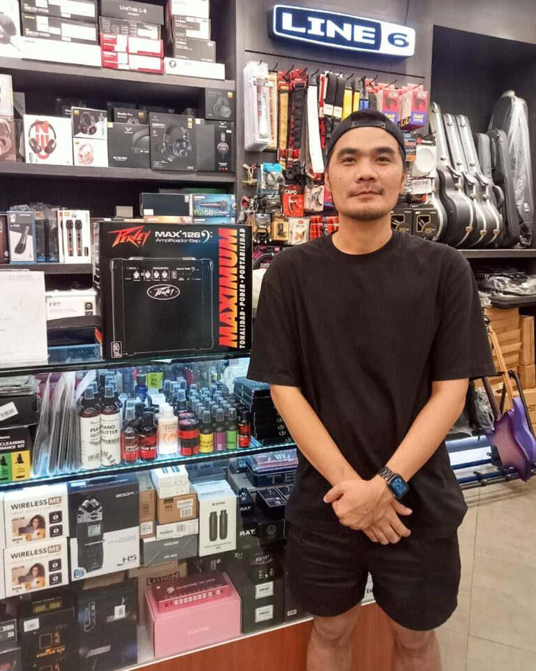 A man in a black shirt and cap stands in a music gear shop, shelves of headphones, guitar cases, and box displays behind him, with a LINE 6 sign above.