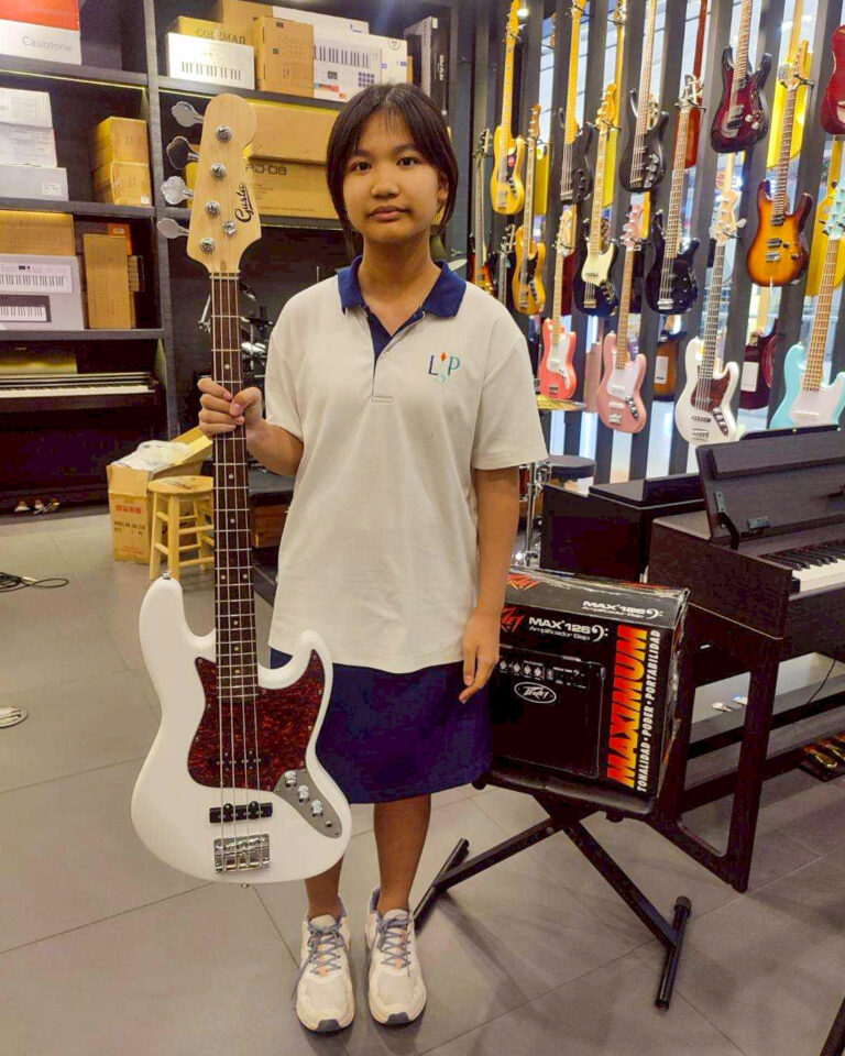 Person standing in a music store holding a white bass guitar with a dark pickguard, with rows of electric guitars on the wall behind her.