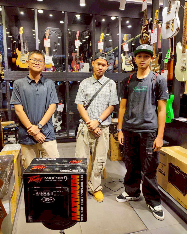 Three young men pose in a guitar shop showroom, with guitars on the wall behind them and amplifiers/boxes on the floor in front of them.