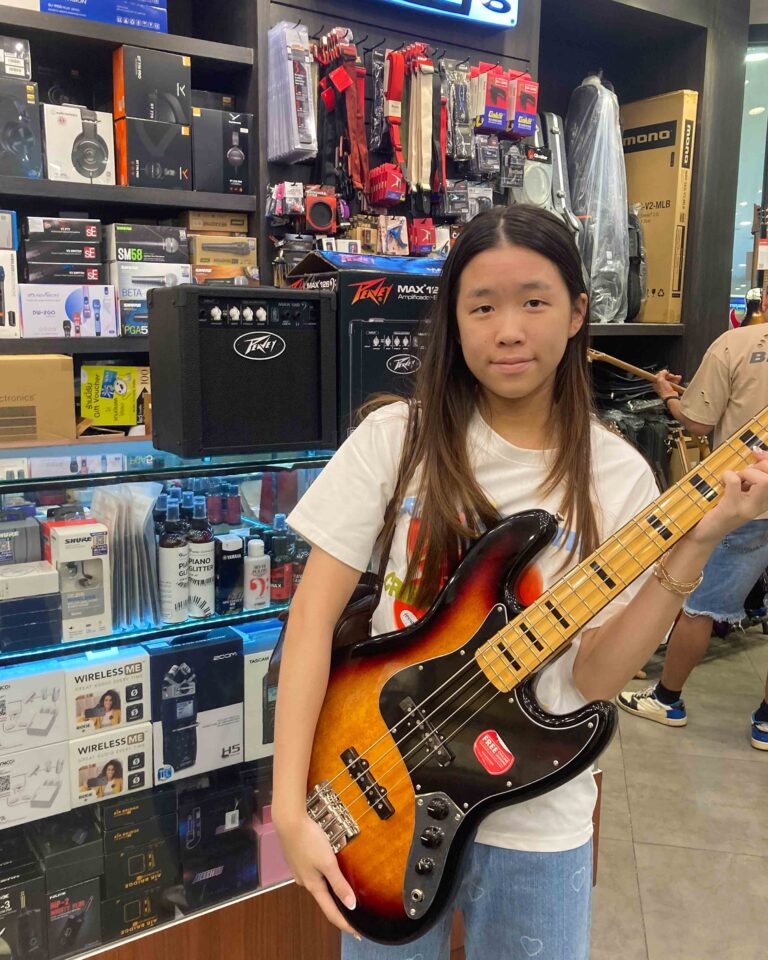 Young woman in a white shirt holding a sunburst bass guitar in a music store with shelves of audio gear behind her