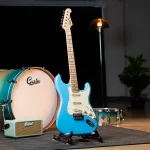 Blue electric guitar on a stand in a music studio, with a turquoise amplifier and a drum kit in the background. ขายราคาพิเศษ