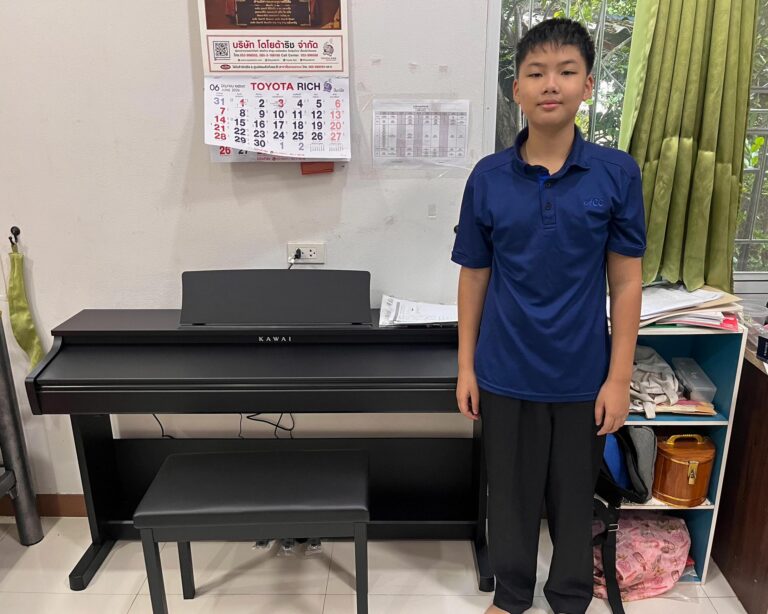 Smiling boy in a blue polo shirt standing beside a black Kawai piano in a room with a calendar on the wall behind him.