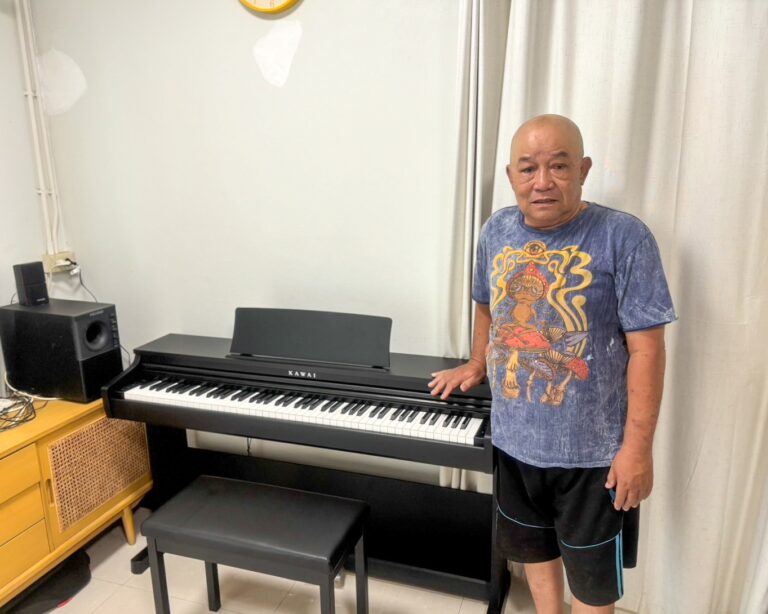 Man in a blue graphic T‑shirt stands beside a black Kawai digital piano in a home setting.